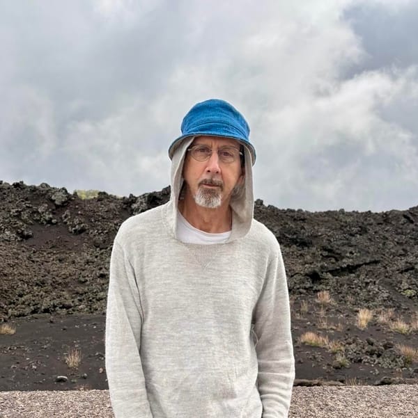 jd hanes, a man with a short mustache and beard wearing glasses, a blue hat, and light grey hoody, stands in front of a rocky